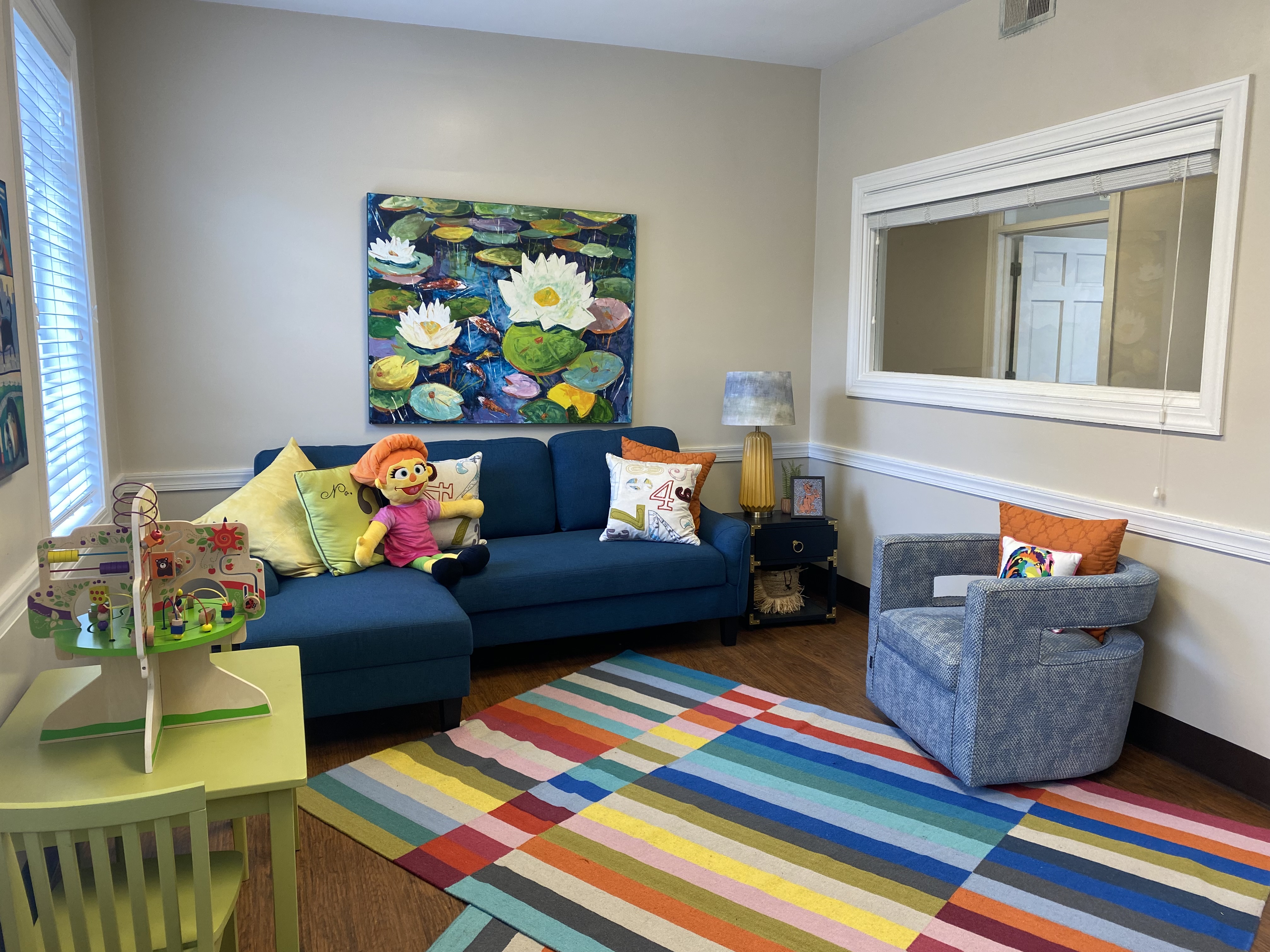 An office at the Center for speaking to children. On the right a half-height window and a round blue chair. On the left a small green table and chair with a children's toy on top, and a blue sectional couch with several throw pillows and a children's toy.