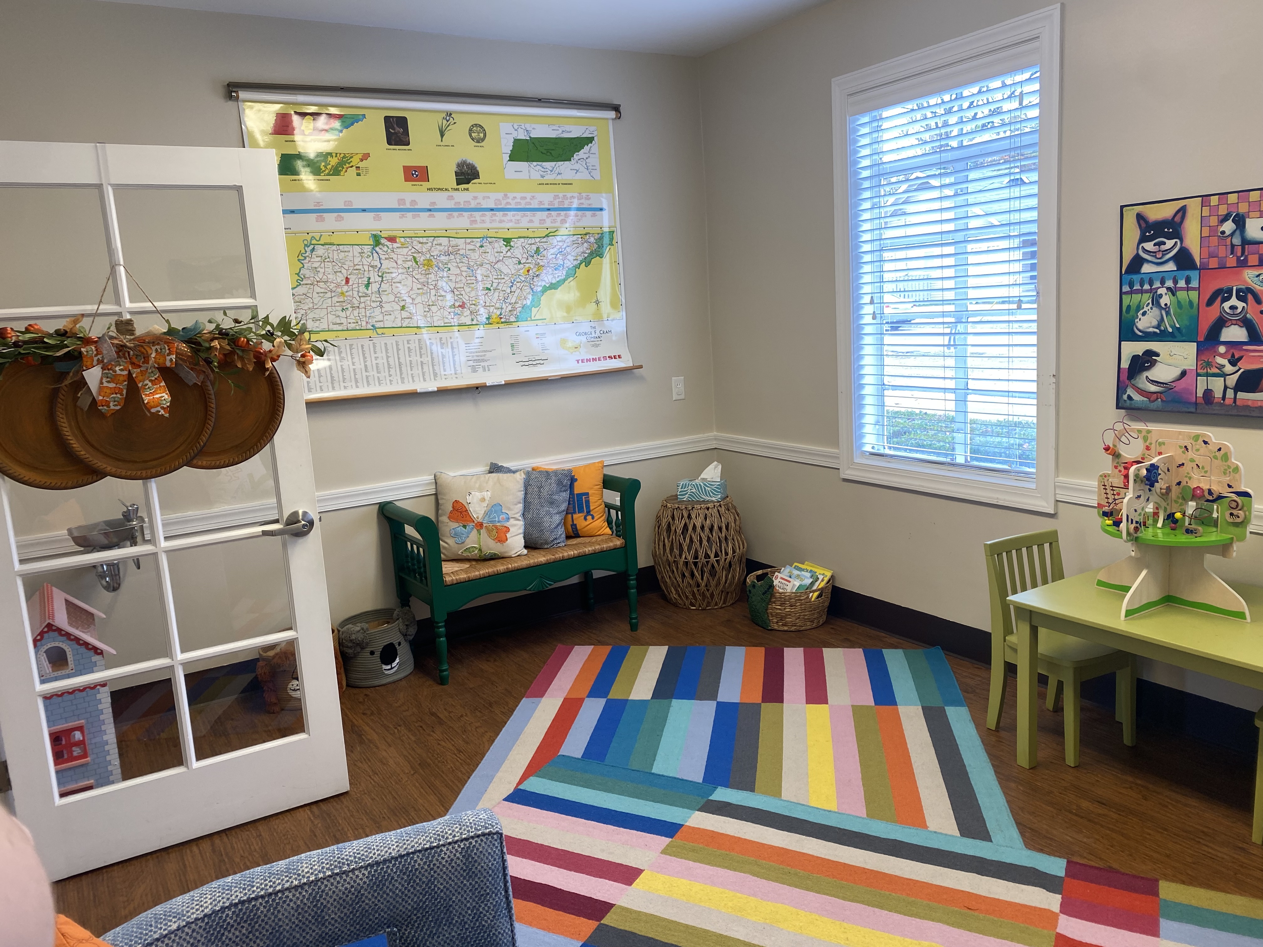 The other side of the room in the previous image. A colorful rug on the floor, the table and chair on the right, an open door with hanging decor, a window on the right side of the wall, and an infographic map of Tennessee on the wall in the center.