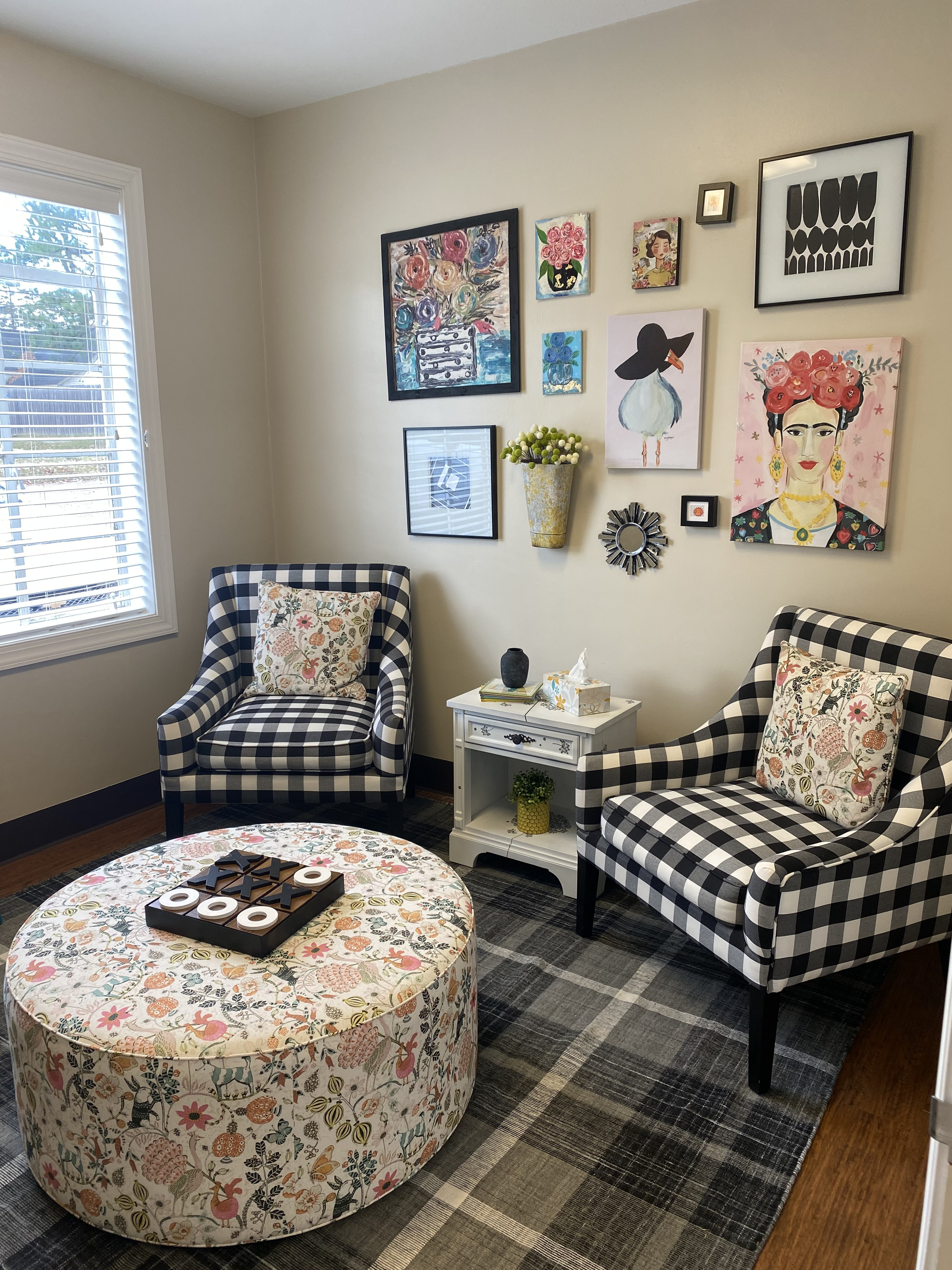 An office at the Center with two matching black and white picnic plaid chairs with matching floral pillows and a large round floral ottoman between the chairs. Several paintings on the wall on the right and a window on the wall on the left.