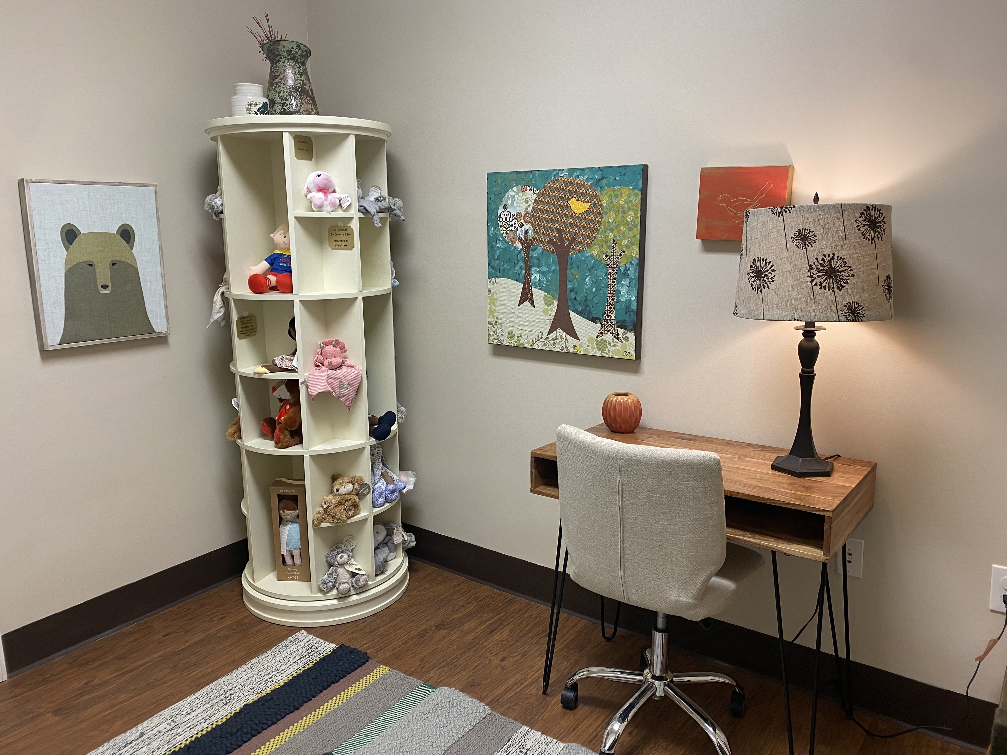 The other side of the room in the previous image, the desk and rolling chair on the right of the image, and in the center a round pillar with cubbies containing stuffed toys and puppets for children. Three paintings on the white wall throughout the image.