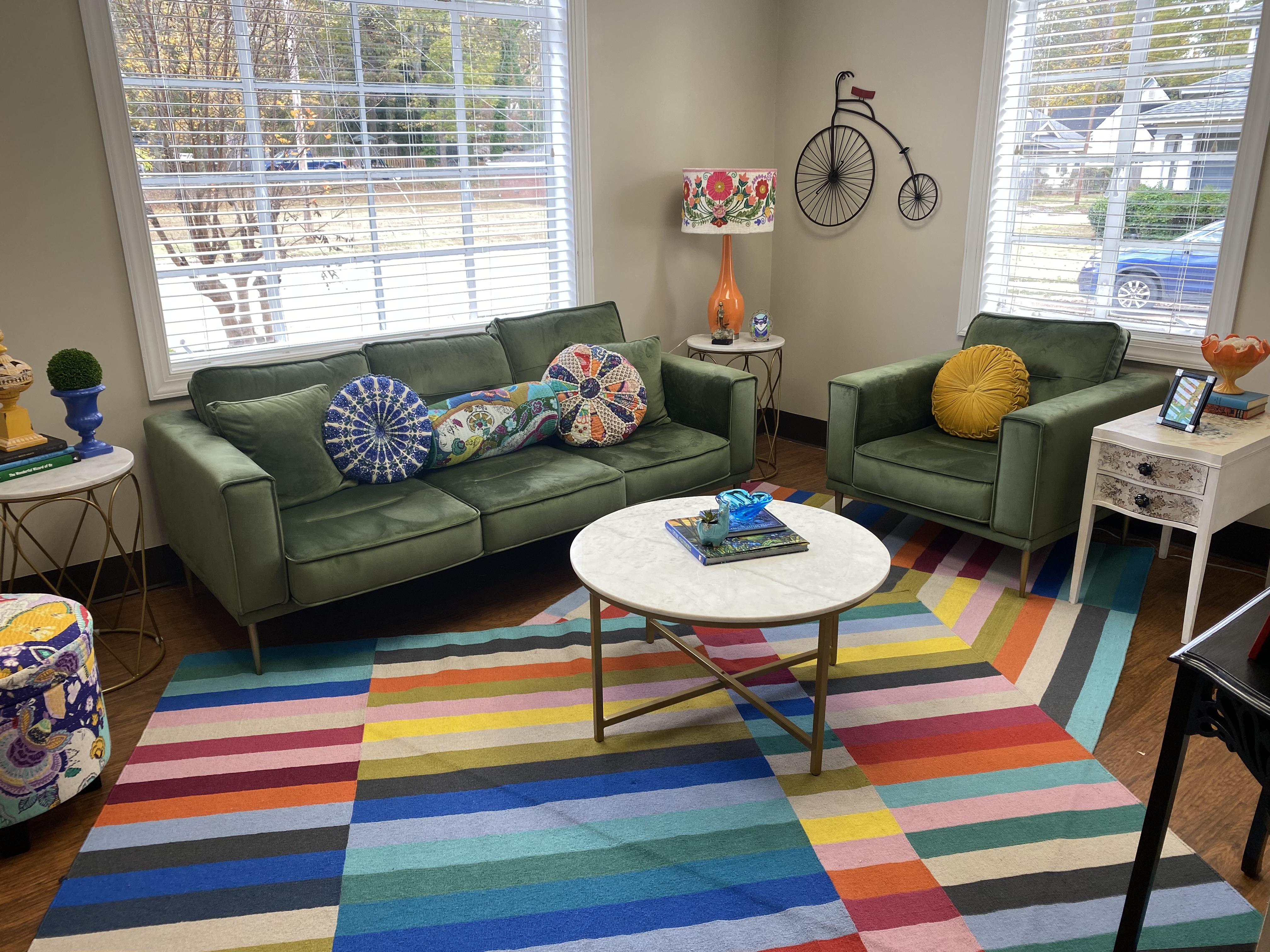 A room at the Center with a green couch, matching green chair, several throw pillows, a colorful rug, and a round white coffee table